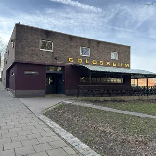 Photograph of a rather plain, boxy brick building. Part of the outside is covered by a green and white veranda. The word 'Colosseum' is written in large, yellow letters on the side of the building.