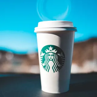 A Starbucks branded paper cup standing on a table and reflecting on the window behind it.